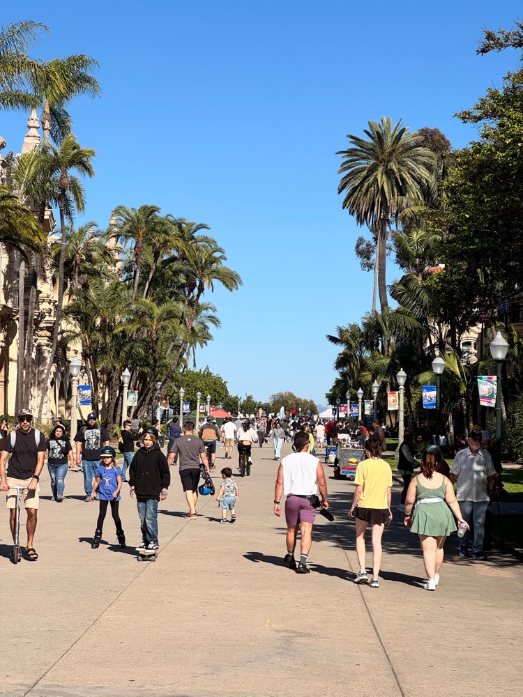 Belebte Promenade im Balboa Park zwischen hohen Palmen und spanischen Kolonialbauten unter blauem Himmel.
