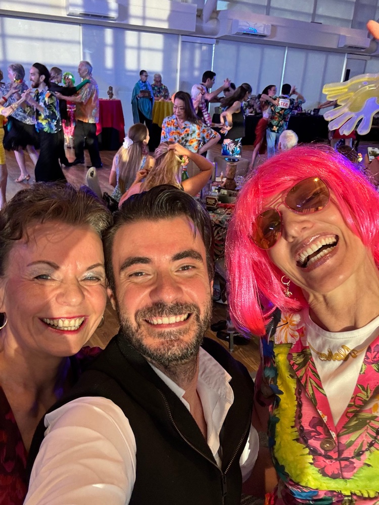 Selfie von drei Personen bei einer Hawaii-Party, rechts eine Frau mit pinker Perücke und Sonnenbrille, dahinter tanzende Paare in Blumenhemden.