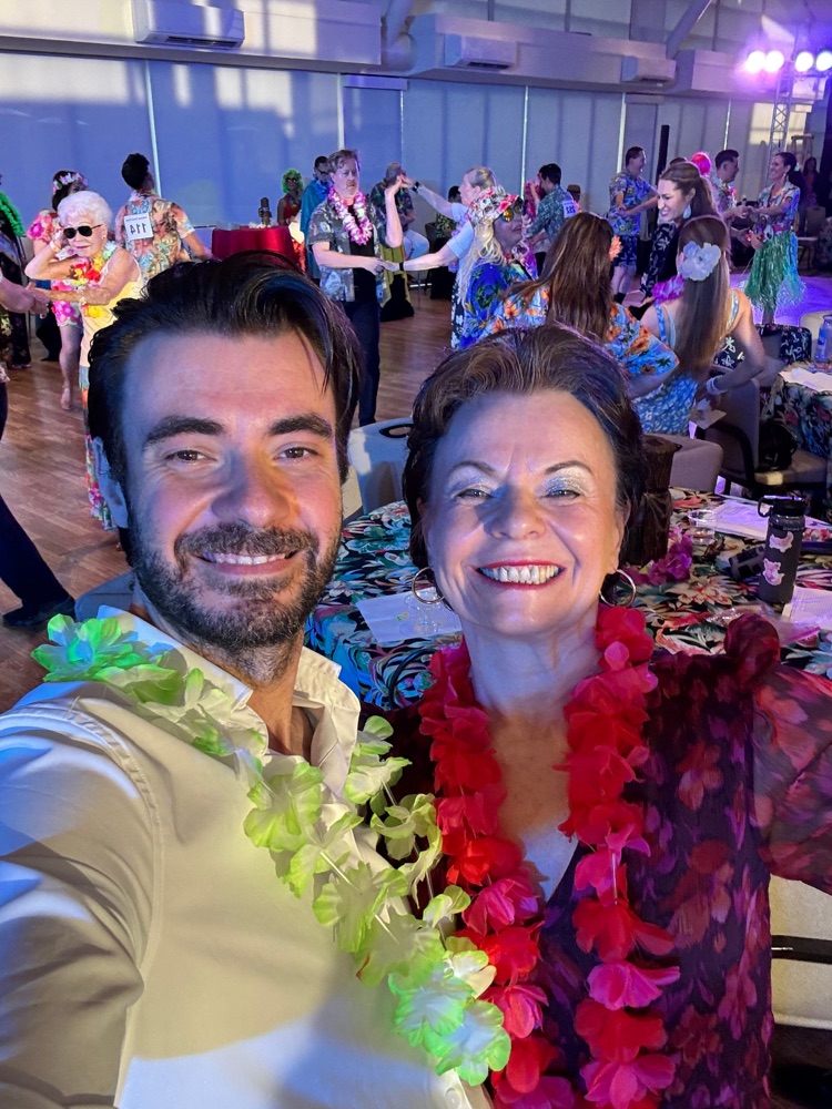 Zwei Personen mit Blumenketten bei einer Party, im Hintergrund Gäste in bunten Hemden.